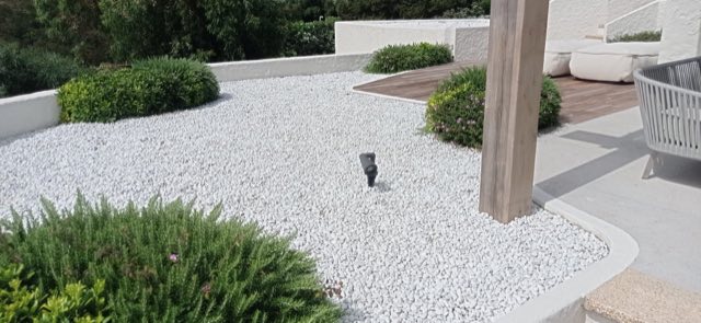 Area in ciottoli bianchi delimitata da arbusti potati a cuscino, accanto a una pedana in legno